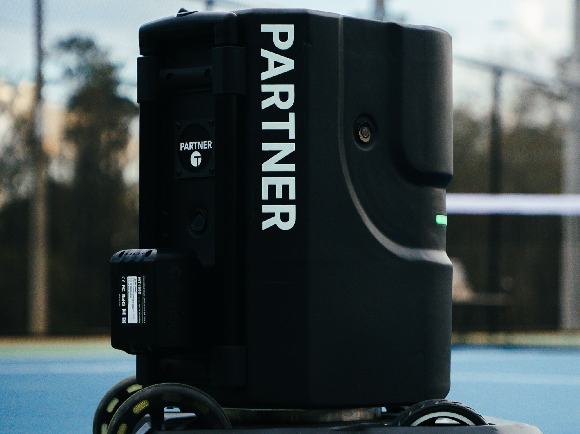 Tennibot Partner V2 tennis ball machine on an outdoor court with the net behind it