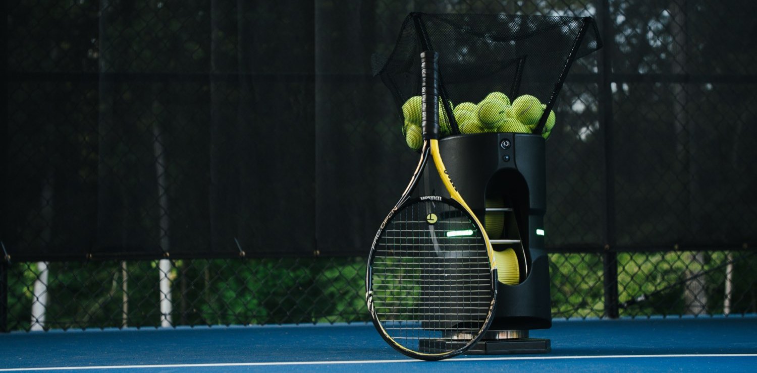 An empty tennis court at dusk with a racquet and a basket of balls at the baseline